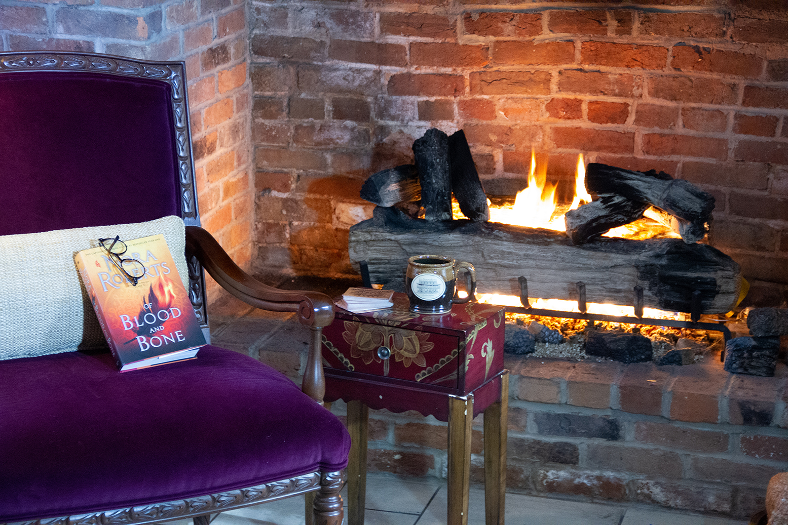 a purple chair and a small table in front of a brick fireplace with a fire burning